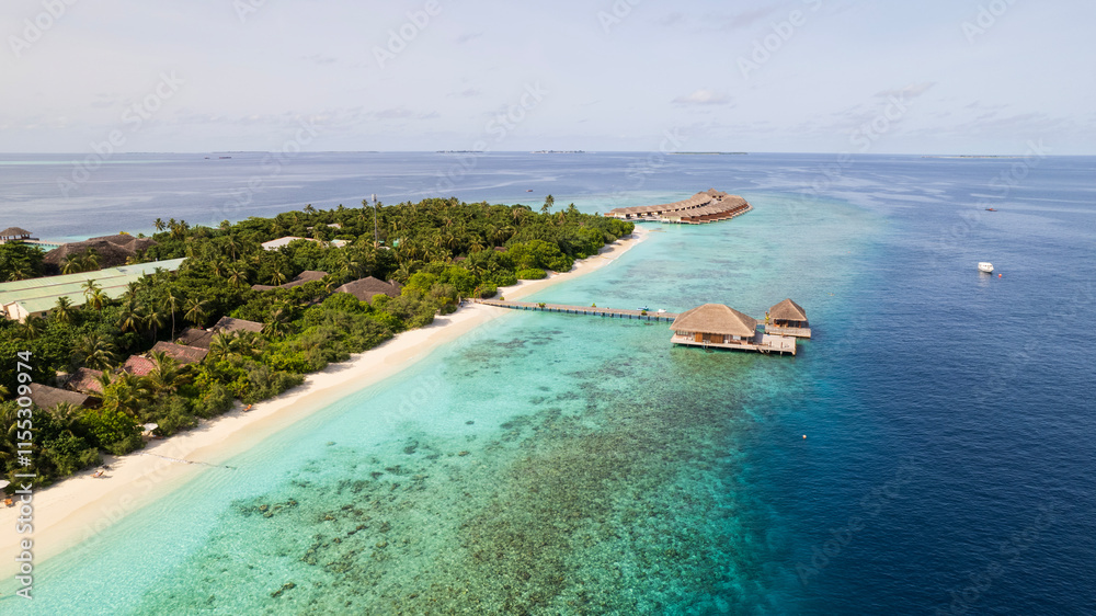 Turquoise clear water and the green island of Maldives. Top view of the ocean and the reef. Palm trees grow on the island. There are bungalows and villas. White sand on the beach. Fish are visible