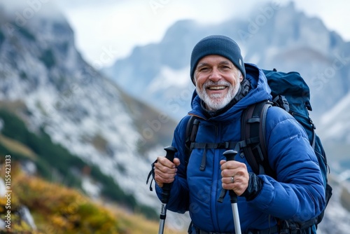 An elderly man clad in a blue jacket and black beanie with hiking poles, stands smiling against a stunning, distant, snow-peaked mountainous background. photo