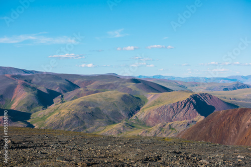 Scenic top view from sunlit stony plain to big colorful mountain range in bright sun under clouds in blue sky. Layered alpine scenery with large multicolor mountains of many vivid colors in sunny day.
