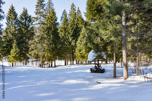 A wooden decorative well covered in snow stands in a snowy forest landscape. Surrounded by evergreen trees under a clear blue sky, this serene winter scene captures the quiet beauty of nature. photo