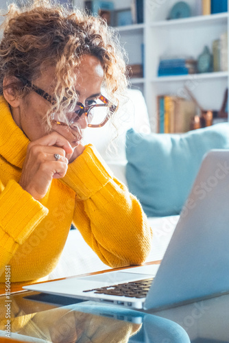 Thoughtful adult woman sitting on her sofa in her living room, concentrating on her laptop for remote work, performing online research, navigating social media, or analyzing work results photo
