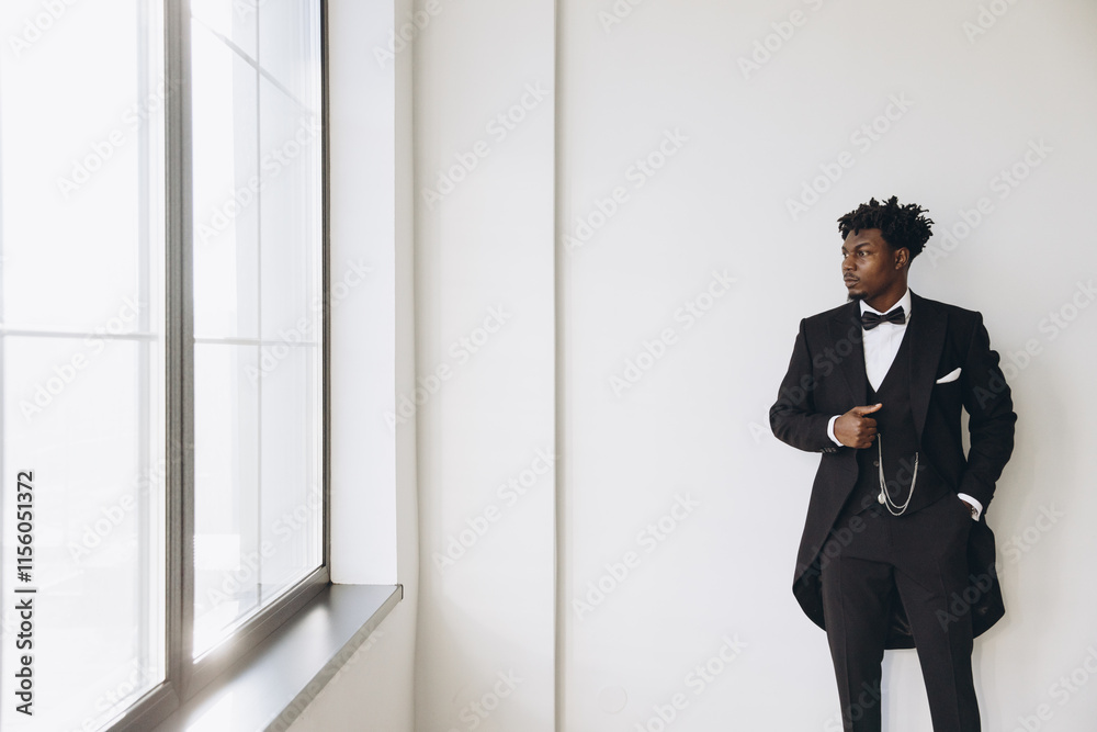 Elegant groom wearing black suit and bow tie posing near window Elegant groom wearing black suit and bow tie posing near window