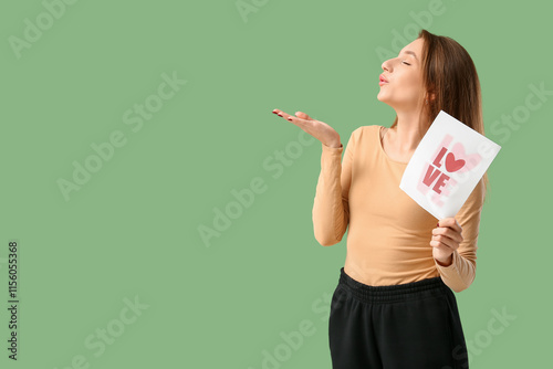 Beautiful young woman holding greeting card with word LOVE and blowing kiss on green background. Valentine's Day celebration