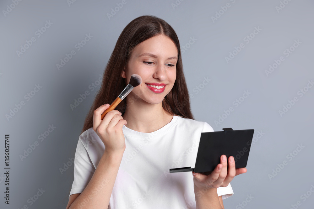 Smiling teenage girl applying blusher with makeup brush on grey background Smiling teenage girl applying blusher with makeup brush on grey background