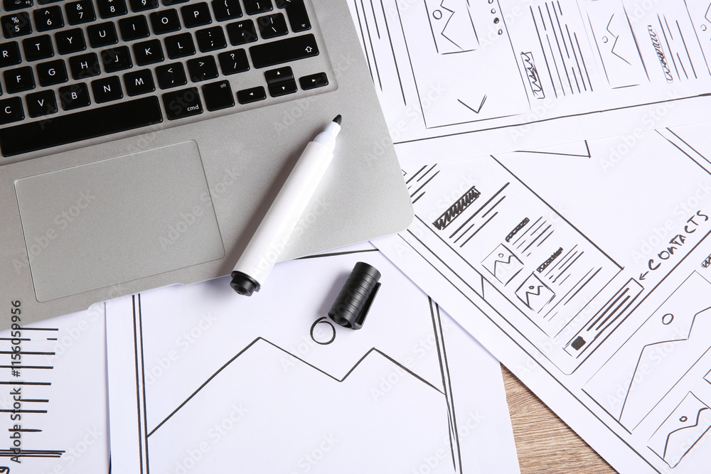 Developing UI design. Sketches of website wireframes, laptop and felt pen on wooden table, above view Developing UI design. Sketches of website wireframes, laptop and felt pen on wooden table, above view