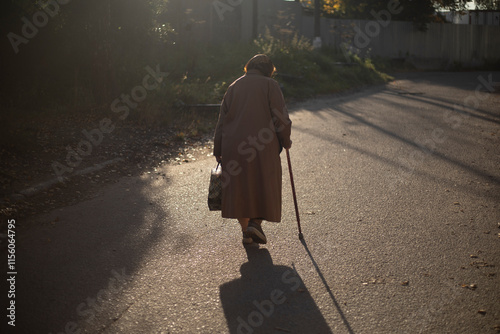 Senior man walking on road in sunlight. Retired woman with walking stick in autumn. An old woman in a coat.