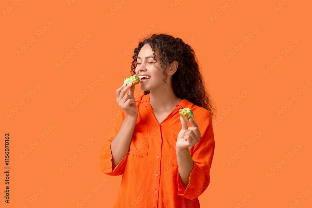 African-American woman eating tasty eclair on orange background African-American woman eating tasty eclair on orange background