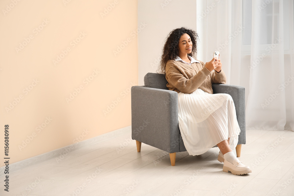 Young African-American woman with mobile phone sitting in grey armchair near beige wall Young African-American woman with mobile phone sitting in grey armchair near beige wall