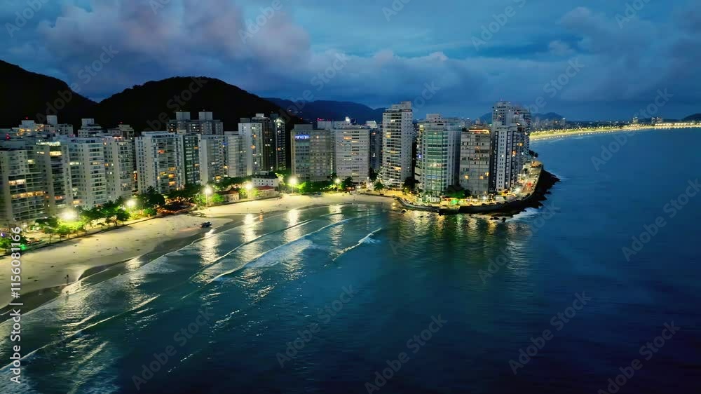 Imagem aérea noturna de praia Pitangueiras no Guarujá com muitas nuvens de chuva e diversos prédios residenciais usados para locação para temporada.
