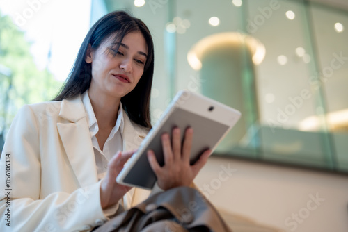 woman in white blazer sits comfortably, using tablet in modern setting. Her focused expression reflects engagement with device, showcasing blend of professionalism and relaxation photo