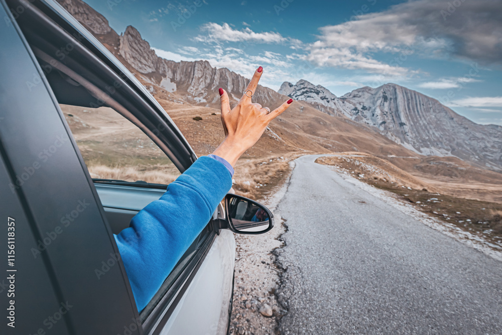 Woman making rock and roll hand gesture from car window while enjoying road trip in the mountains