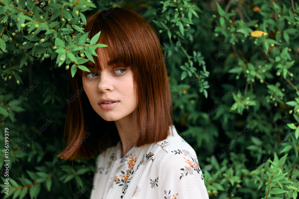 Mysterious woman with red hair standing in front of lush green plants and bushes gazes off to the side Mysterious woman with red hair standing in front of lush green plants and bushes gazes off to the side