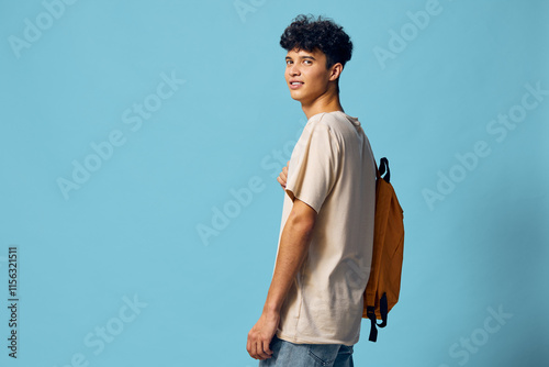 Young man smiling against a blue background, wearing a casual beige t shirt and holding a yellow backpack, reflecting a relaxed and youthful vibe in an urban setting photo