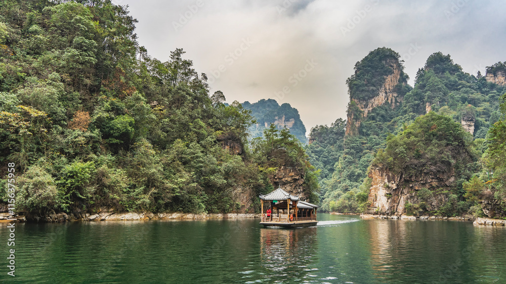 Obraz premium A beautiful Chinese boat with curved tiled roofs, canopies and carved wooden railings floats on a emerald lake. A man in a life jacket is on board. Green vegetation on coastal hills and cliffs. China.