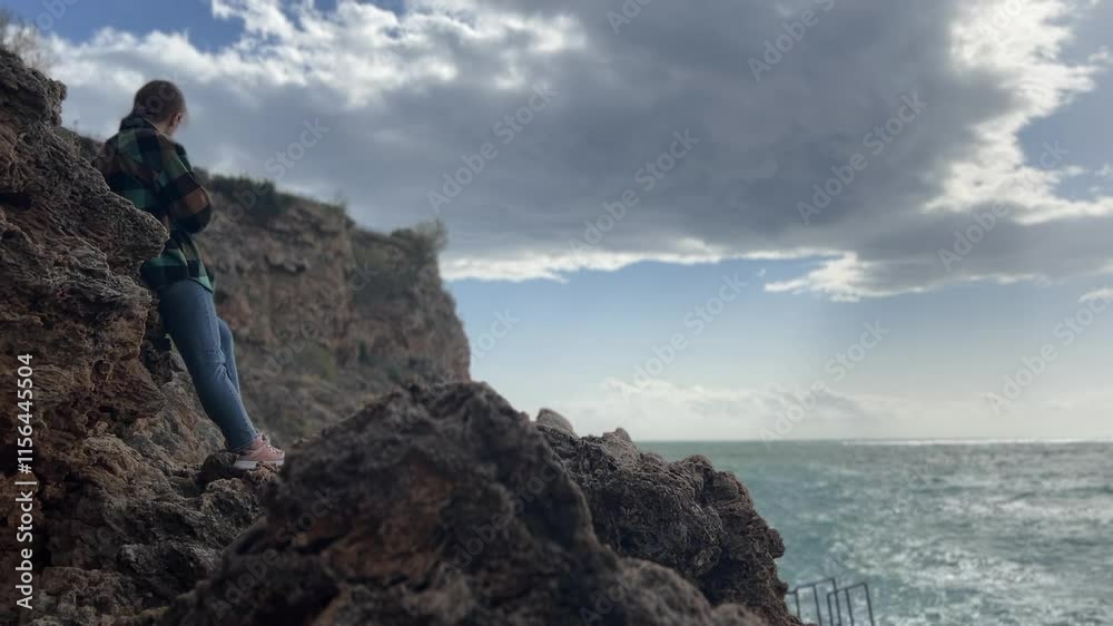 A solitary woman leans against jagged rocks, her expression reflective as she looks out over the ocean, with a windy sky full of dramatic clouds above. A solitary woman leans against jagged rocks, her expression reflective as she looks out over the ocean, with a windy sky full of dramatic clouds above.