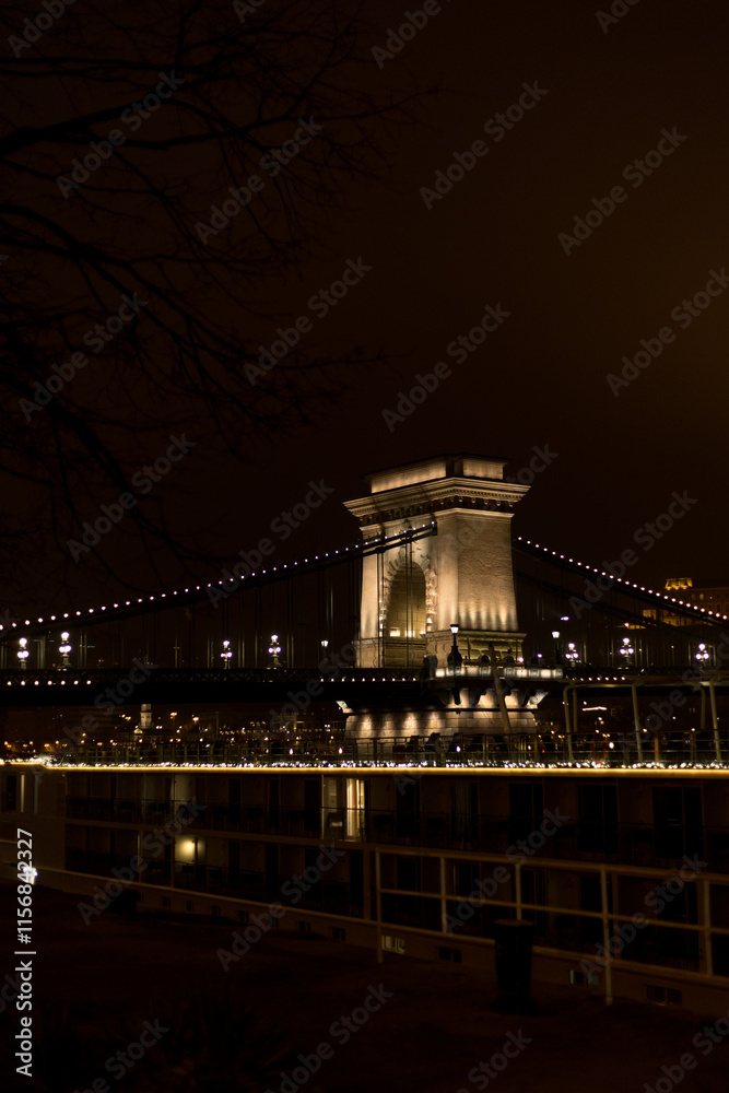 Obraz premium A captivating nighttime view of the iconic Chain Bridge in Budapest, Hungary. The bridge glows warmly under city lights, connecting the two sides of the Danube River.