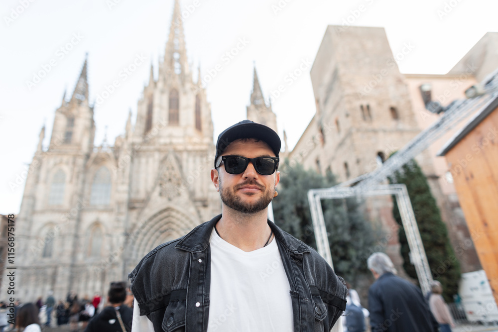 Obraz premium Handsome traveler cool man with sunglasses with cap in denim vest walking near church in Barcelona, Spain
