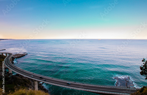 A breathtaking view of a curving coastal bridge over the ocean under a bright blue sky, surrounded by pristine waters and rugged cliffs. The horizon stretches endlessly into the distance