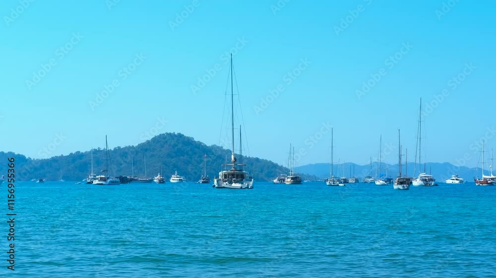 Sailboats gently swaying on calm turquoise water. The boats are moored near a hilly shore on a sunny summer day
