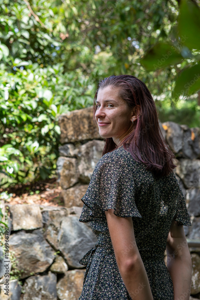 Fototapeta premium A smiling woman in a floral dress stands outdoors in front of stone steps surrounded by lush greenery. The natural light and vibrant backdrop enhance the cheerful and serene atmosphere