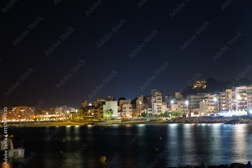 People relaxing on beach at night, watching summer festival with illuminated buildings in background