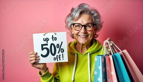 Smiling senior woman holds shopping bags, sign showing 50 off. Happy elderly shopper displays holiday sale items. In studio setting against pink background. Black Friday promotion. Discounts on photo