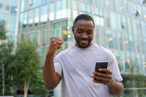 Happy African American man euphoric winner with smartphone on street in city. Person guy looking at cell phone reading great news getting good result winning online bid feeling amazed. Winning gesture