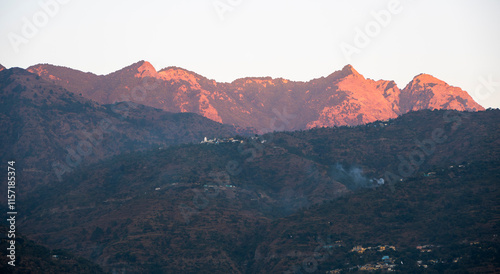 The sunset casts a warm glow over the mountains, with a small town visible on the lower hill. Smoke arising from the houses adds to the charm of Tehri Lake. photo