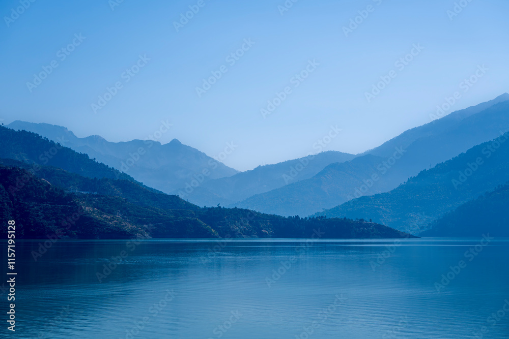 Multilayered blue mountains are shrouded in mist, with blue light cascading over them and the lake waters. This picturesque scene at Tehri Lake captures the enchanting interplay of light and nature. Multilayered blue mountains are shrouded in mist, with blue light cascading over them and the lake waters. This picturesque scene at Tehri Lake captures the enchanting interplay of light and nature.