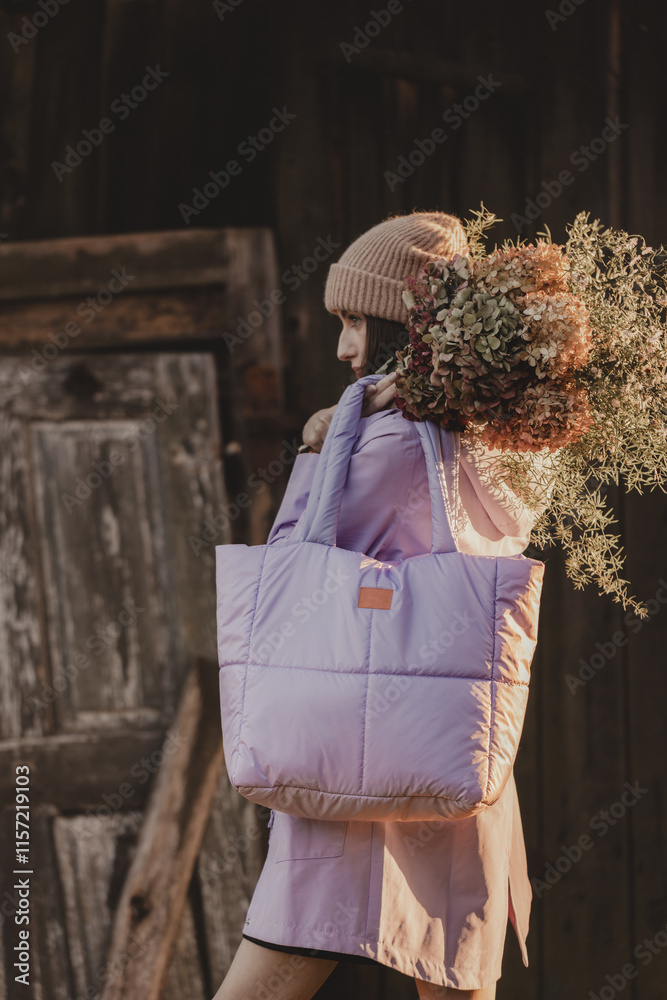 Naklejka premium A woman in a hat and a light purple jacket with a large quilted bag and a bouquet of dried flowers poses in front of a wooden wall in subdued light (selective focus)