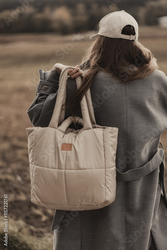 A woman in a gray coat with her back inverted back holding a beige bag against the background of the field (selective focus)