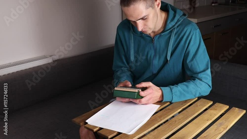 man in blue hoodie tunes green kalimba with handwritten tablature on paper nearby. The scene captures playing instrument.playing musical instrument with your thumbs photo