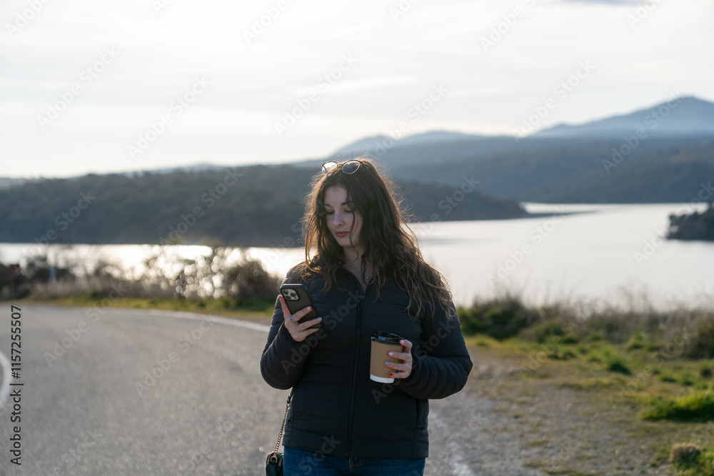 Young woman using smartphone and drinking coffee by the lake on a road trip Young woman using smartphone and drinking coffee by the lake on a road trip