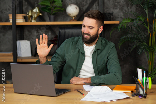 Young happy man freelancer or entrepreneur have video conference, shows a greeting gesture, working remotely online at office.