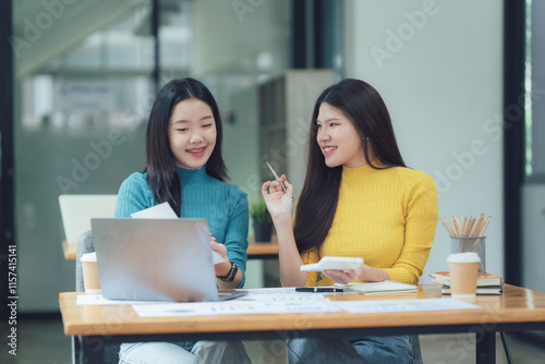 Creative Collaboration: Two young Asian female designers brainstorm ideas, using a laptop and sketches in a modern workspace.  Their smiles reflect a positive and productive atmosphere.  photo