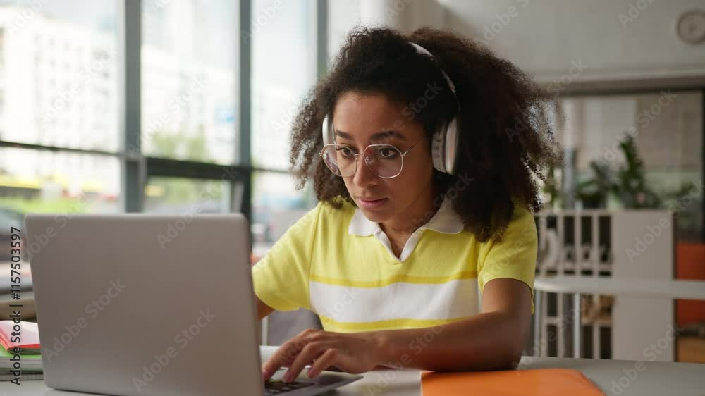 African American woman student girl ethnic female wearing headphones writing homework prepare exams test studying online video class lesson internet with laptop indoors school university e-learning