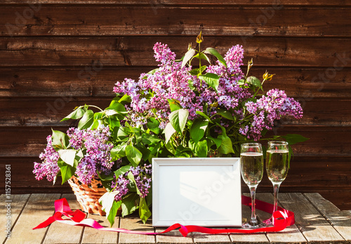 White frame, glasses of champagne and  branches of lilac in vase and in wicker basket on aged rustic wooden table in spring sunny day. View with copy space.