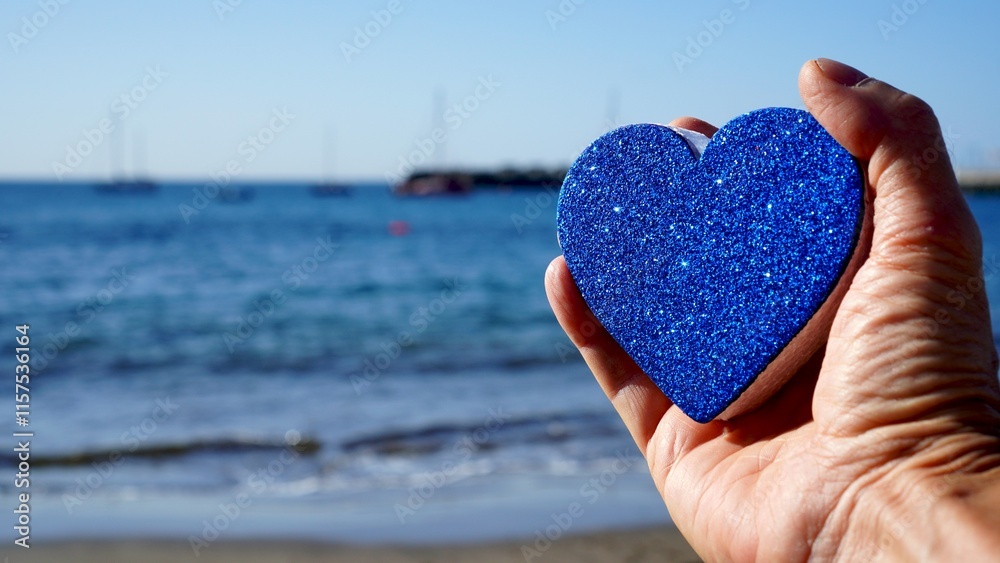 Fototapeta premium Valentines day seaside. Hand holds heart with blue glitter in front of the sea. Sailboats on horizon in the blurred background. Love and romance concepts