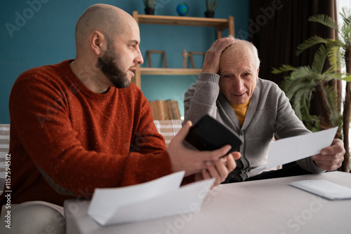 Adult son helping old father pay bills count expenses using calculator sitting on couch at home photo