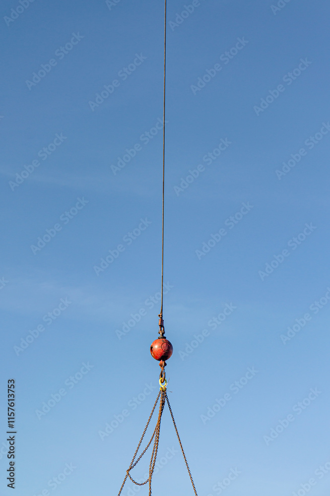 Front view, far distance of, an industrial crane's red ball weight, under load, hanging from crain chains with light blue-sky background Front view, far distance of, an industrial crane's red ball weight, under load, hanging from crain chains with light blue-sky background