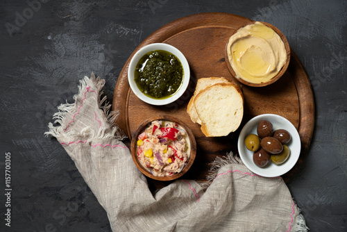 Light breakfast in small bowls on a wooden round board: pesto, tuna salad, hummus, olives and fresh baguette. A perfect moment to enjoy with space for text.
 photo