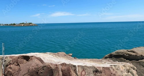 4K drone footage of a sunny seascape with rocky shores and a distant lighthouse. A stunning coastal view ideal for travel, summer, and nature-related projects.