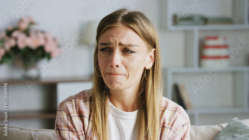 Fotografie Young woman sitting on a comfortable sofa at home is crying and wiping tears fro