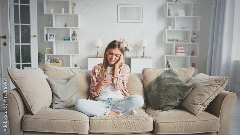 Obraz premium Young woman sitting on a comfortable sofa at home, reading bad news or receiving upsetting message on smartphone and feeling worried, stressed, frustrated, or tired