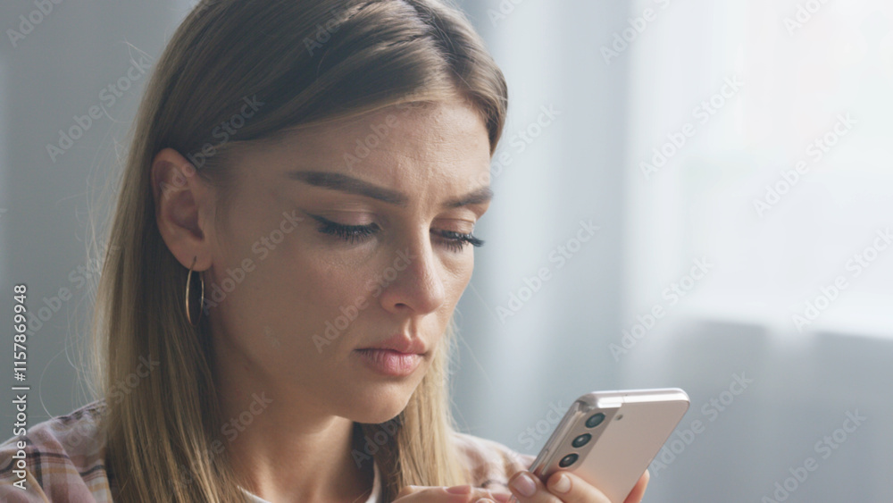 Young woman with long blond hair and earrings holding mobile phone and reading message with worried facial expression, standing next to window in bright room Young woman with long blond hair and earrings holding mobile phone and reading message with worried facial expression, standing next to window in bright room
