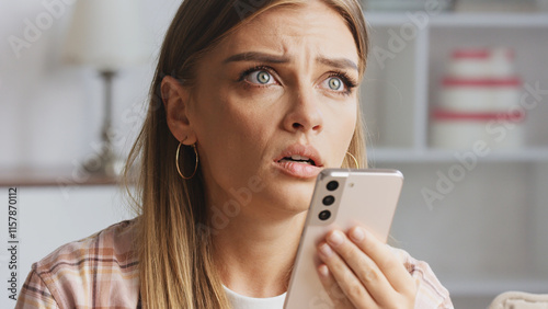 Young woman with long blond hair and gold hoop earrings holding a rose gold smartphone, displaying a shocked expression while listening to a surprising voice message indoors photo