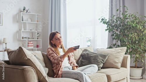 Bored woman sitting on a comfortable sofa in her living room, holding a remote control and changing channels while watching television, expressing disinterest and a lack of engaging content