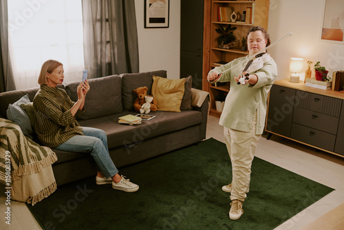 Wide shot of senior woman holding smartphone recording video of her daughter with Down syndrome playing violin in cozy living room photo