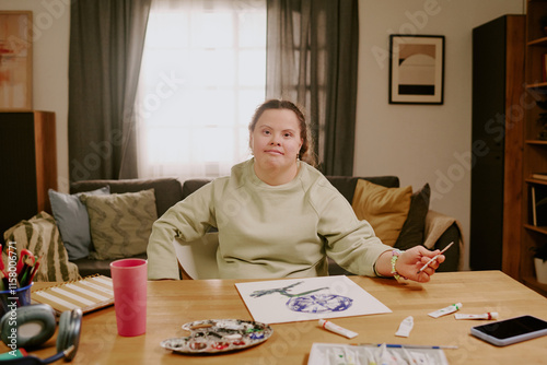 Medium portrait of young girl with Down syndrome sitting at desk in cozy living room, looking at camera, while drawing picture on paper photo