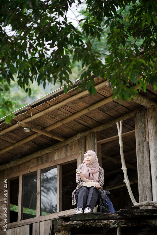 Fototapeta premium young Indonesian woman in hijab reading a book while enjoying nature in a forest cabin, travel concept.